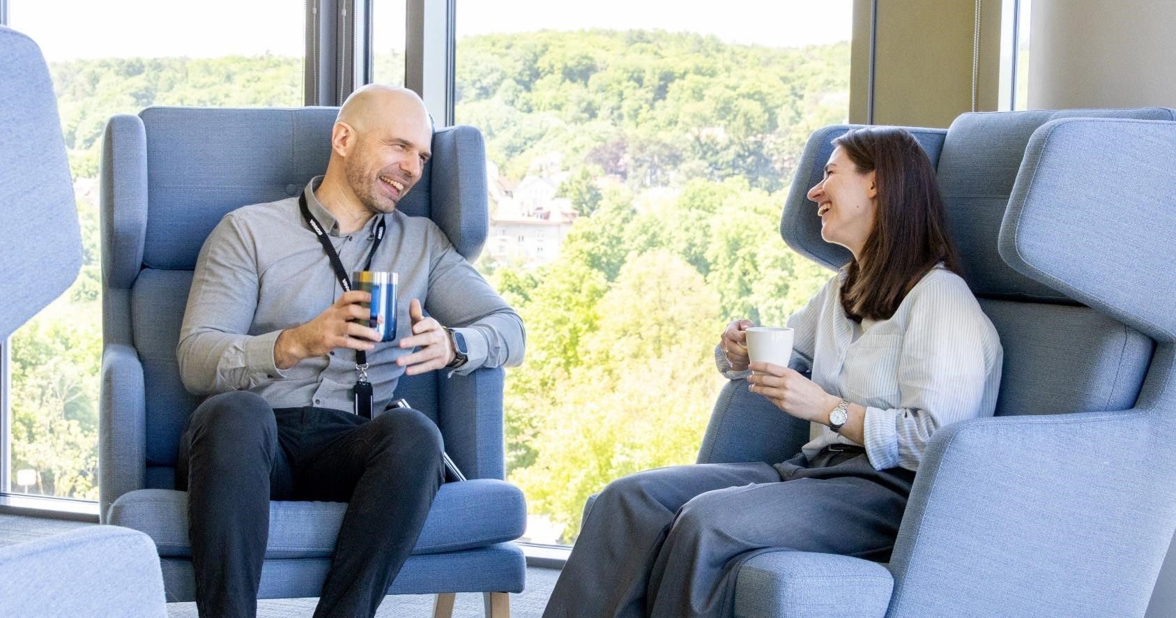 Two Scania employees sitting down in office, talking, laughing and drinking coffee