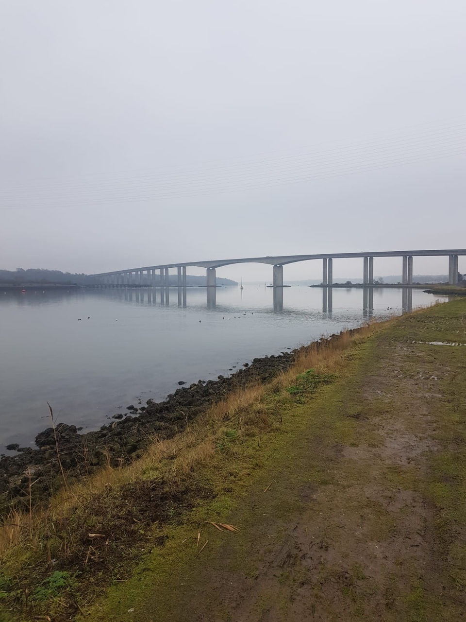 Photo of pathway next to river with view of bridge in fog