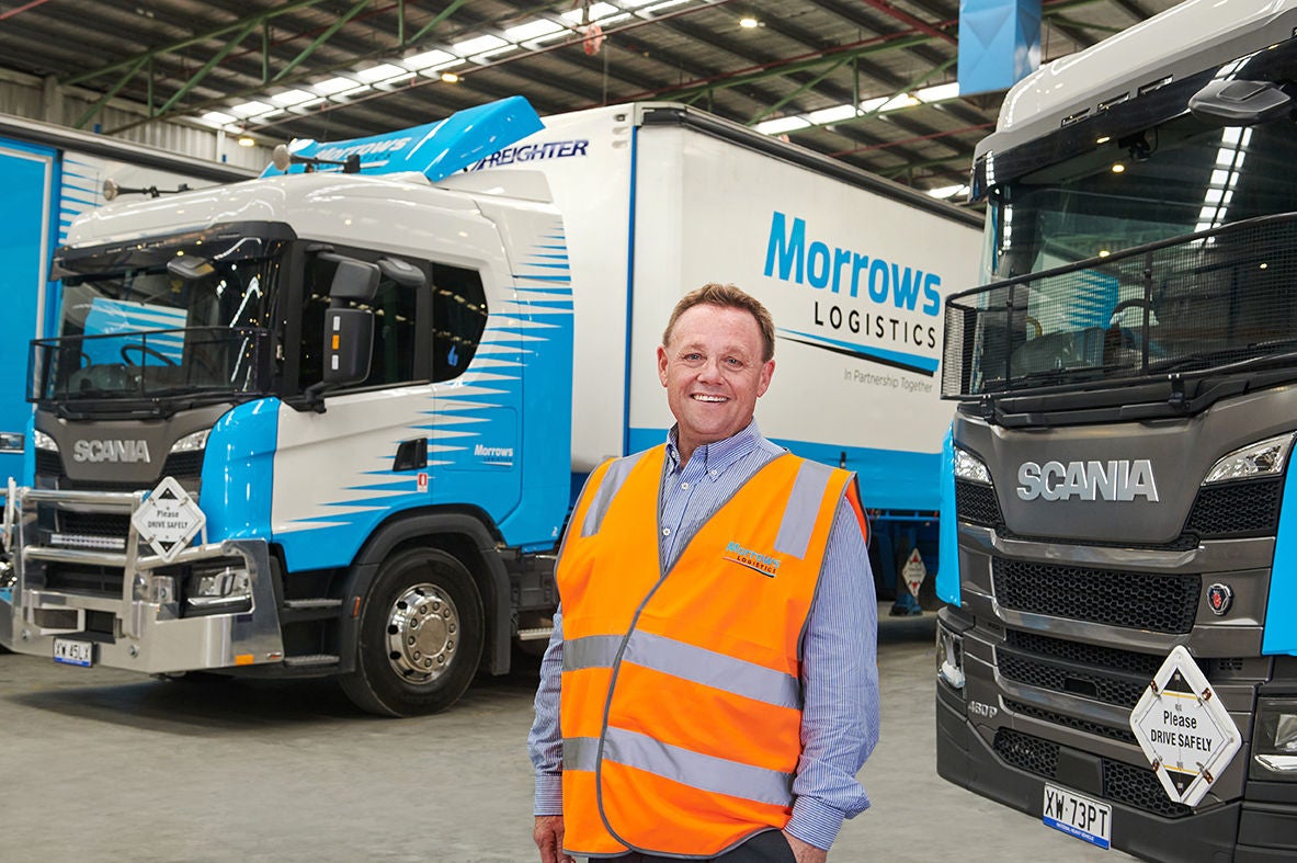 Man in an orange hi-vis vest standing in a warehouse between two Scania trucks branded with the Morrows Logistics livery.