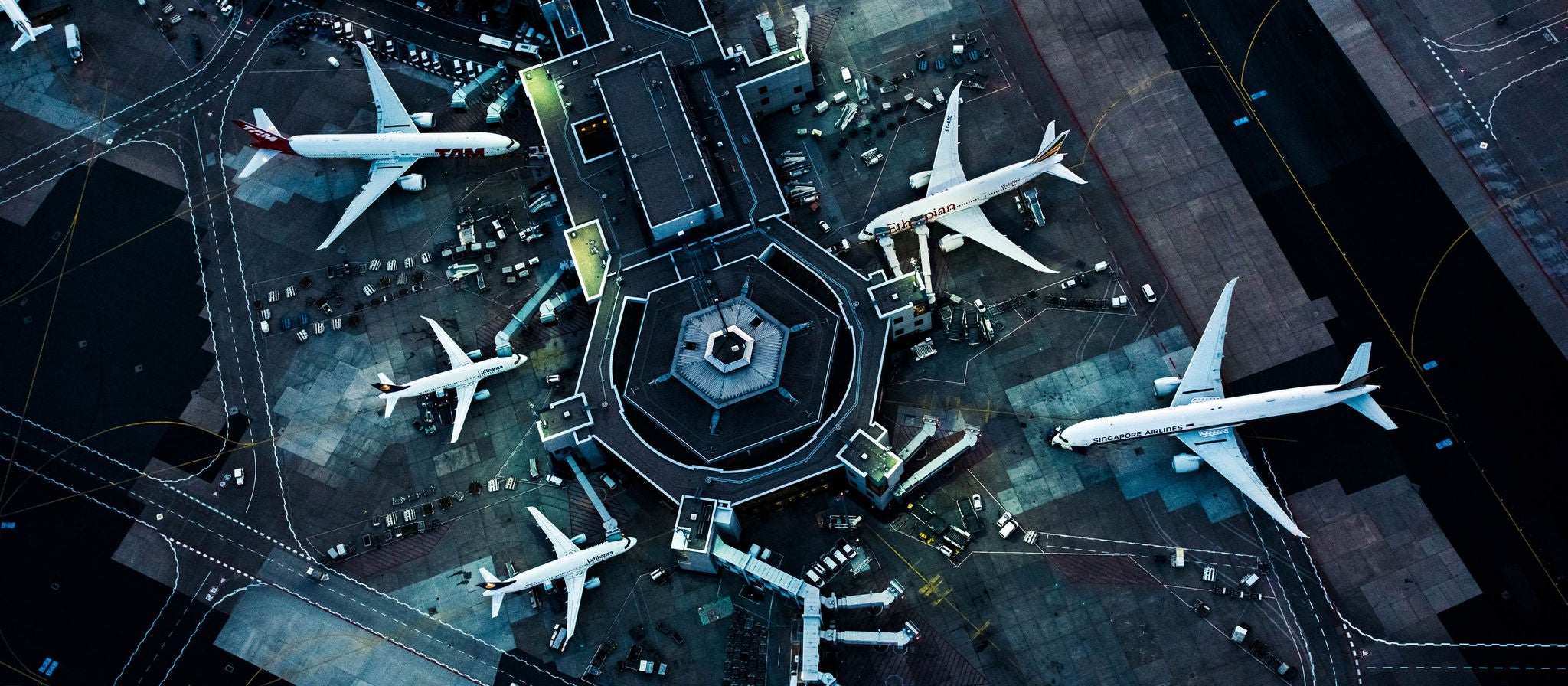 Aerial view of an airport terminal with five passenger aircraft parked at gates, surrounded by service vehicles and jet bridges.