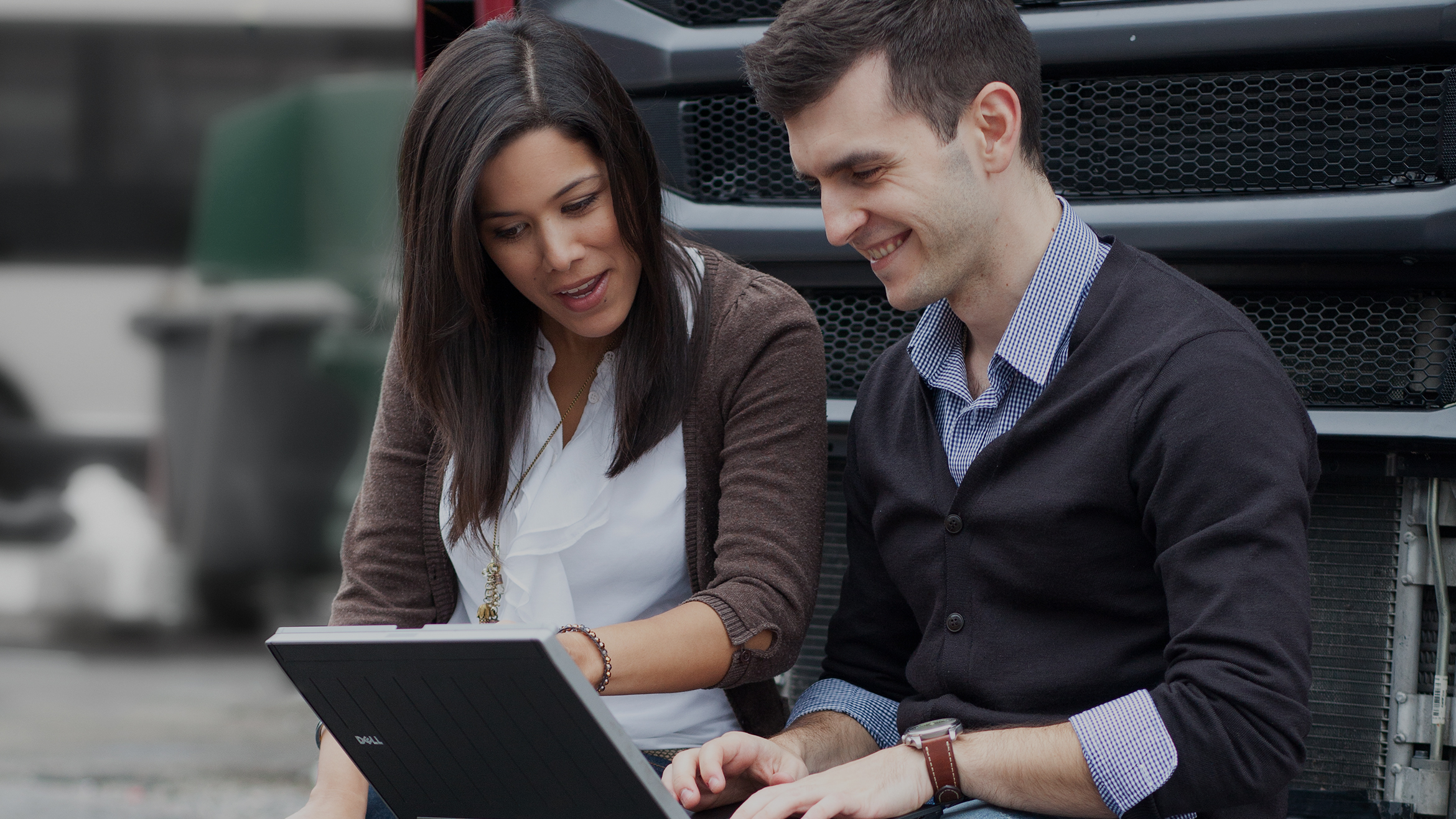 Two employees leaning against a truck looking at a laptop Two employees leaning against a truck looking at a laptop