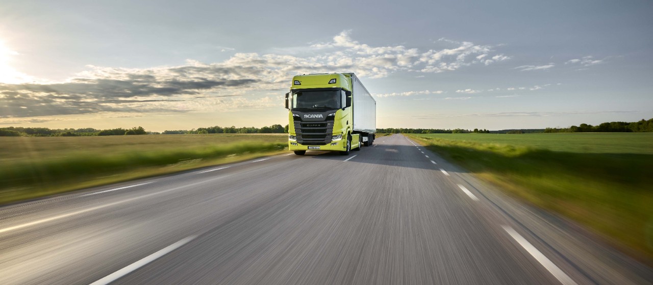 Een gele Scania-vrachtwagen op een weg die door groene velden wordt doorkruist