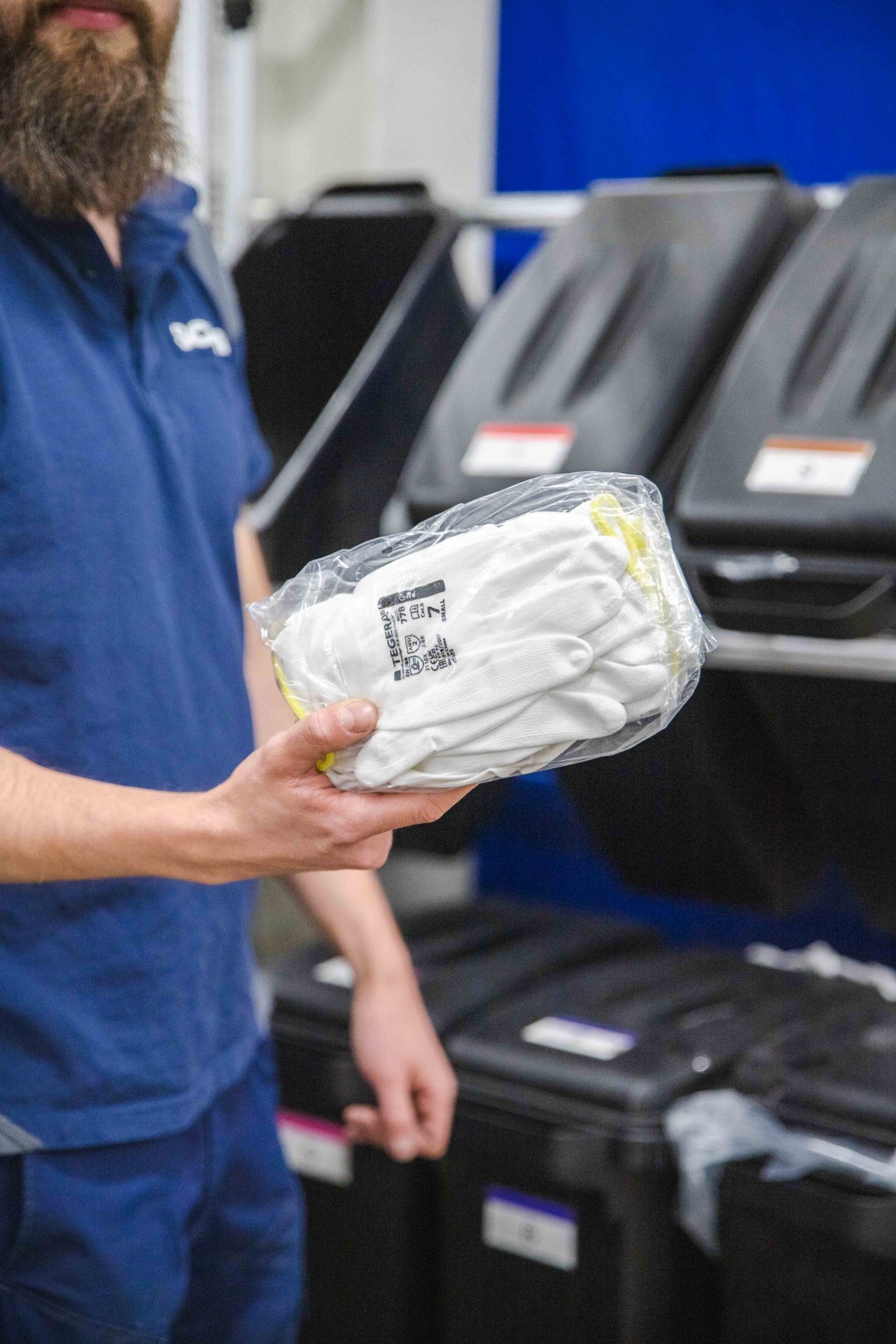 Medarbetare håller upp en genomskinlig plastförpackning innehållandes vita skyddshandskar framför återvinningskärl