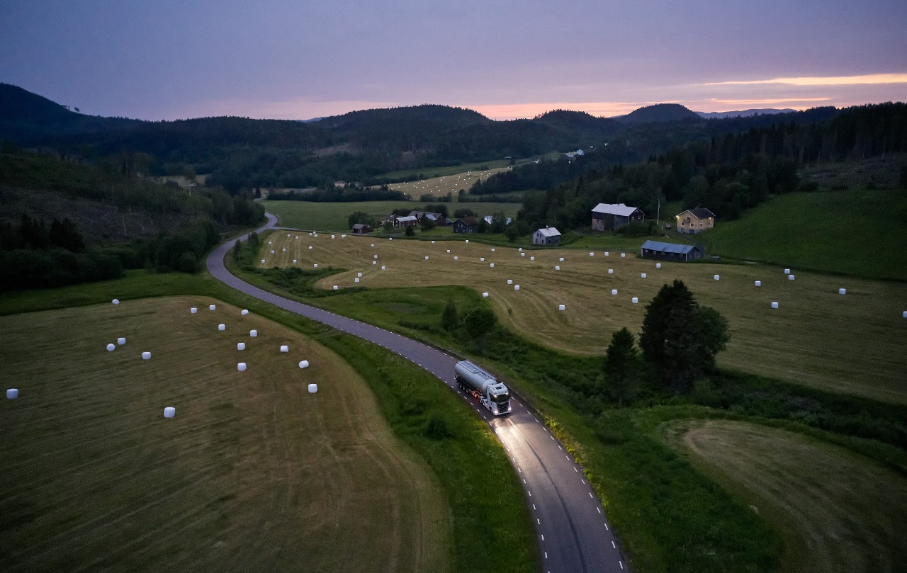 En Scania-lastbil som åker längs med en väg med åkrar på båda sidor i solnedgång