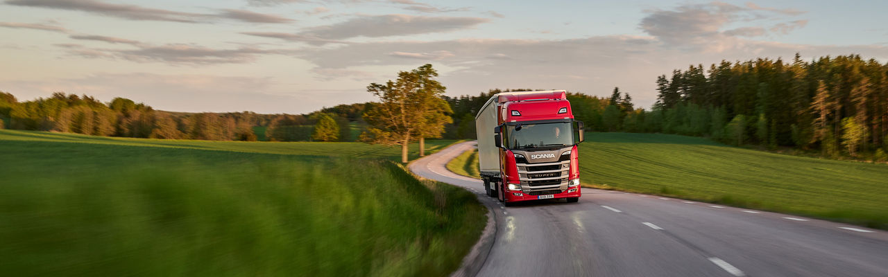En röd Scanialastbil på en väg som omges av gröna fält